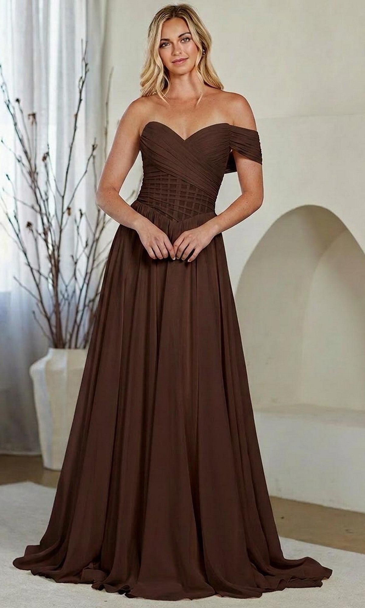 A woman stands indoors near a white wall and decorative branches, wearing the Amelia Couture 9097 long brown prom dress featuring a strapless, pleated bodice and off-the-shoulder sleeves.